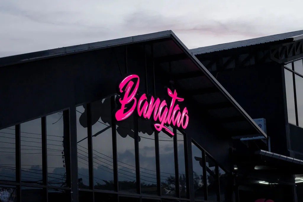 The glass-fronted Bangtao Muay Thai & MMA training building at dusk, marked by its pink neon sign.
